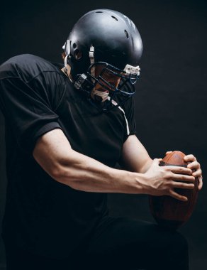 Kasklı ve zırhlı Amerikan futbolcu siyah duvara karşı top taşıyor.