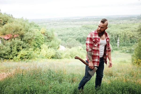 Skäggig timmerarbetare stående i skogen med yxa — Stockfoto