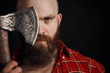 Bıyıklı bir adam savaş baltası tutuyor. Gözünü kapatıyor ve siyah arka plandaki kameraya bakıyor.