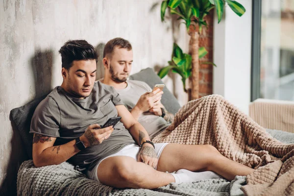 Feliz pareja gay acostados juntos en la cama y el uso de teléfono inteligente, mirando las fotos — Foto de Stock