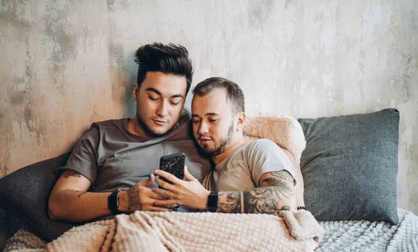 Boldog meleg pár feküdt együtt az ágyban, és használja okostelefon, nézi a képeket — Stock Fotó