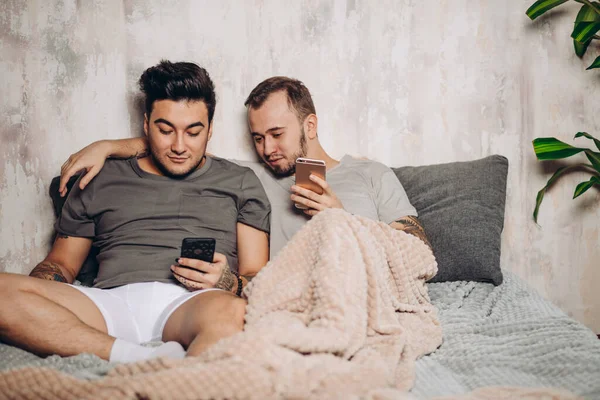 Felice coppia gay sdraiato insieme a letto e utilizzando smartphone, guardando le foto — Foto Stock