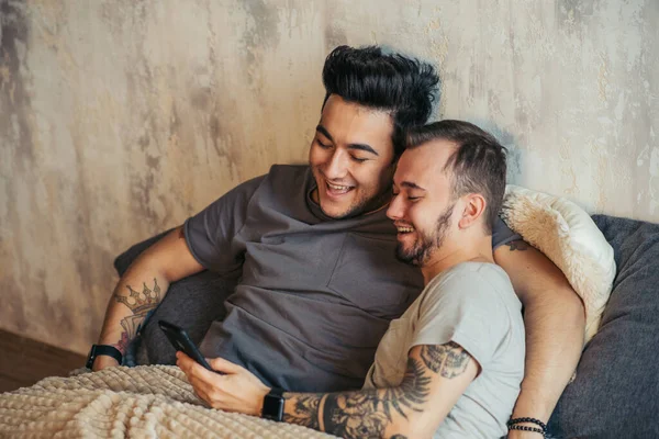 Feliz pareja gay acostados juntos en la cama y el uso de teléfono inteligente, mirando las fotos —  Fotos de Stock