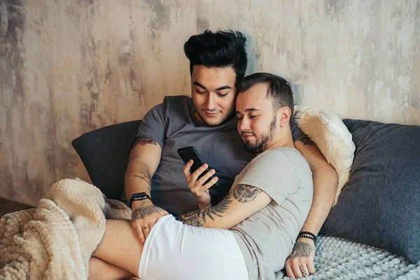 Heureux gay couple couché ensemble dans lit et en utilisant smartphone regarder photos — Photo