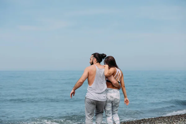 Joven pareja de fitness abrazándose de pie en la playa, mirando al horizonte —  Fotos de Stock