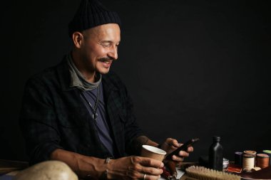 Deri ustası akıllı telefon kullanıyor ve elinde bir fincan kahve tutuyor.