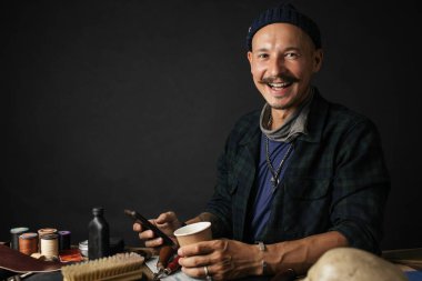 Deri ustası akıllı telefon kullanıyor ve elinde bir fincan kahve tutuyor.