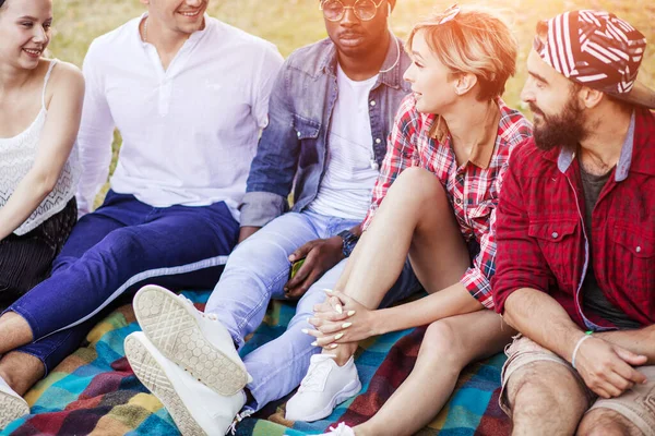 Diverse groep vrolijke vrienden zittend op gras bij zonsondergang in natuurpark. — Stockfoto