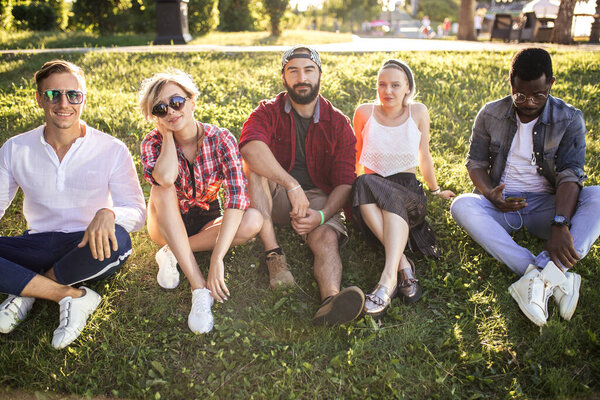 Diverse group of cheerful friends sitting on grass at sunset in nature park.