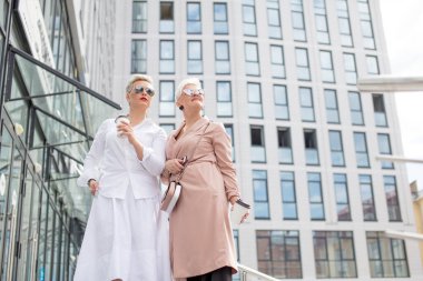 İki iş kadını arka planda binaları olan bir kahveyle dışarıda duruyor.