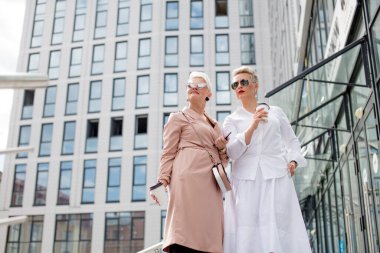 İki iş kadını arka planda binaları olan bir kahveyle dışarıda duruyor.