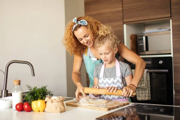 Hermosa mamá joven y su linda hijita rodando la masa en casa. — Foto de Stock