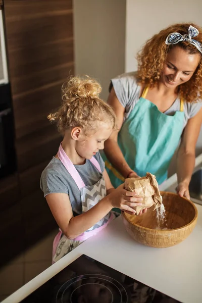 Niña y su madre rociando harina en un tazón y sonriendo mientras hornean — Foto de Stock