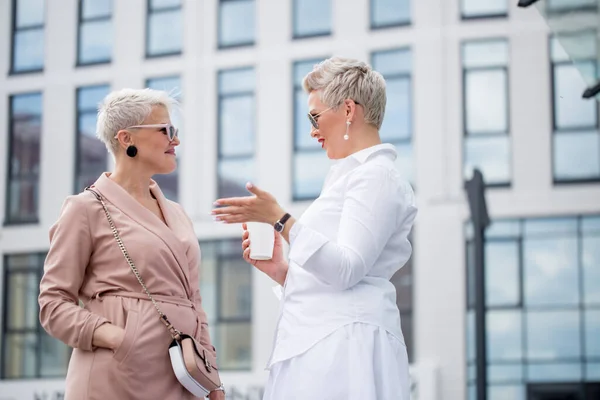 two businesswomen standing outside with coffee with buildings on background