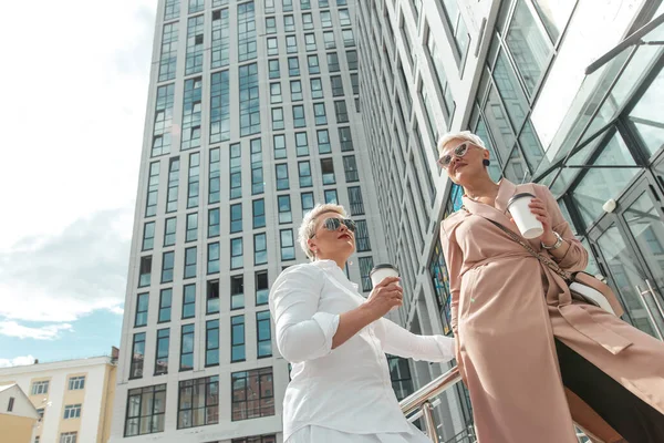 Dos mujeres de negocios de pie afuera con café con edificios en el fondo —  Fotos de Stock