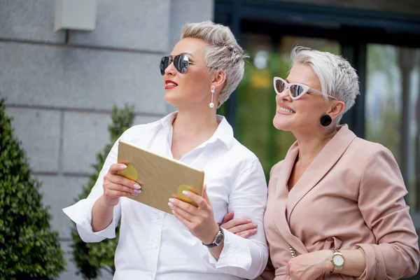 Dos empresarias caminando por la calle cerca del edificio. mujer de negocios que va junto con la tableta —  Fotos de Stock