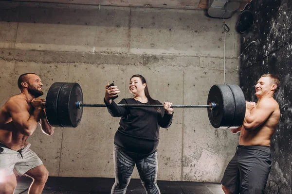 Crazy fat girl taking a picture of herself while sportsmen lifting the barbell — Stock Photo, Image