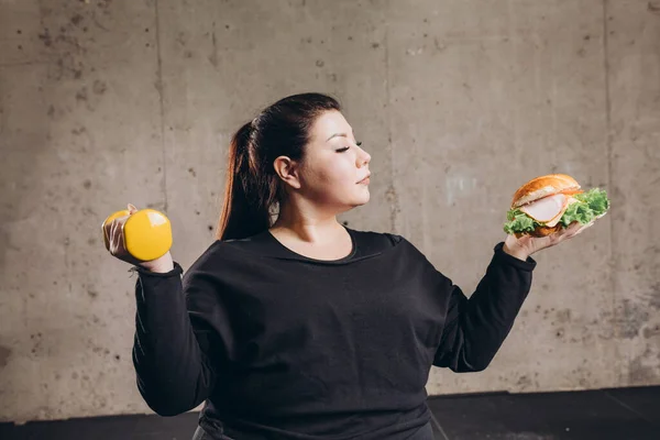 Otyła kobieta trzyma hantle i hamburgery, odmawiając uprawiania sportu, uzależnienia — Zdjęcie stockowe
