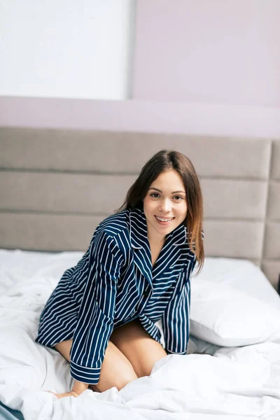 Chica atractiva positiva en camisa de moda a rayas sentada en la cama —  Fotos de Stock