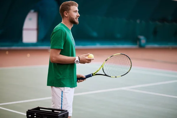 Hombre barbudo joven en ropa deportiva verde está jugando al tenis en la cancha cubierta. — Foto de Stock