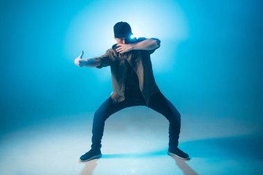 İzole edilmiş neon mavi arka planda hip hop dansı yapan genç bir dansçı.