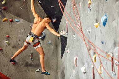 Erkek tırmanışçı spor salonunda egzersiz yapıyor, yapay duvara tırmanırken kayalar kullanıyor..