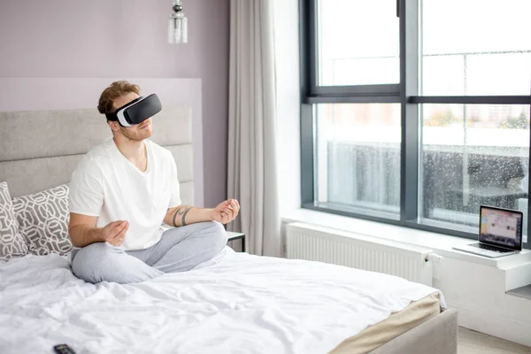 Agradable hombre en VR auriculares sentado en postura de yoga en la cama —  Fotos de Stock