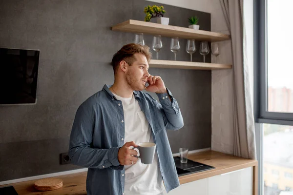 Junger gutaussehender Mann, der zu Hause Kaffee trinkt und auf dem Smartphone spricht — Stockfoto