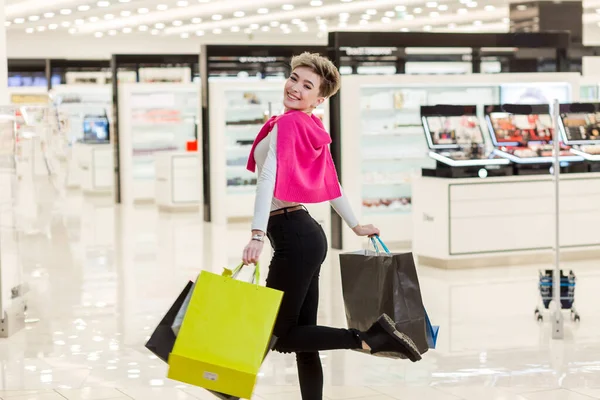 Glimlachende vrouw draagt een aantal boodschappentassen met winkels op de achtergrond — Stockfoto