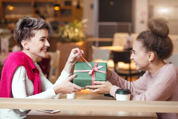 Mujer feliz felicita a su novia con la caja de regalo mientras está sentado en la cafetería. —  Fotos de Stock