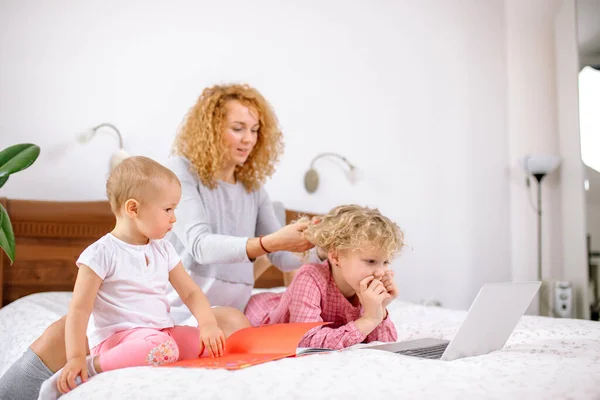 Mother brushing her daughters hair while resting on the bed with a baby — Stock Photo, Image