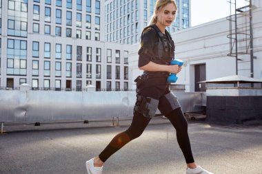 Sarı saçlı kız elinde halterlerle sokakta koşuyor.