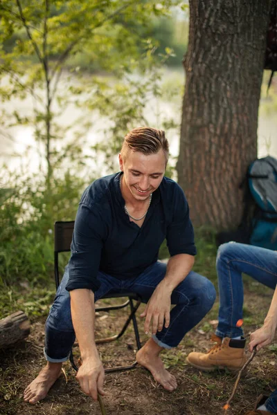 Atraktivní muž v košili a džínách sedí kolem táboráku — Stock fotografie