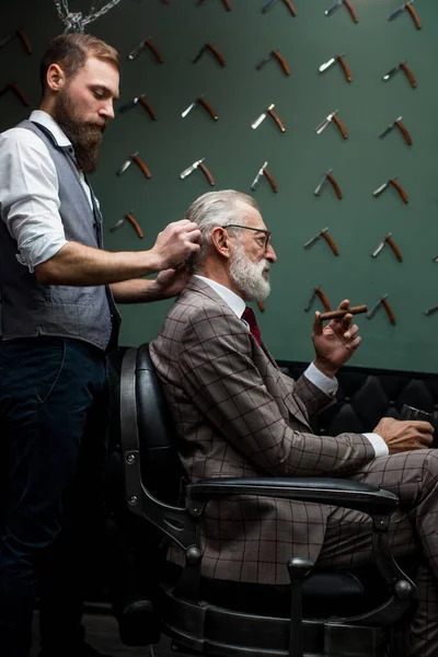 Bearded old businessman sitting at barber shop in chair choosing haircut design