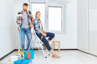 Gülümseyen bir çift evde resim yapıyor, ellerinde boya silindiri ve fırça tutuyorlar.