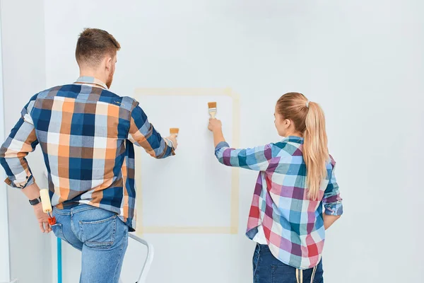 Couple in casual plaid shirts paint house wall together. DIY renovation concept. — Stock Photo, Image