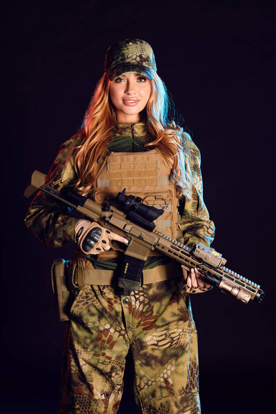 Sniper woman in ghillie suit, cap, plate carrier holds airsoft rifle in studio