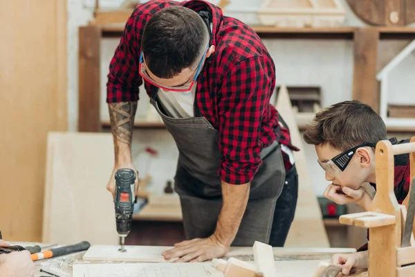 Father and little son working with drill perforating wood plank at workshop — Stock Photo, Image