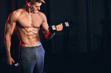 Kas gövdesi olan erkek antrenör, spor salonunda elinde dambıllar tutuyor.