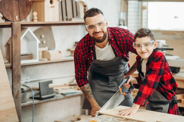 Papá y su hijo están trabajando en el taller. Están aserrando tablones de madera . — Foto de Stock