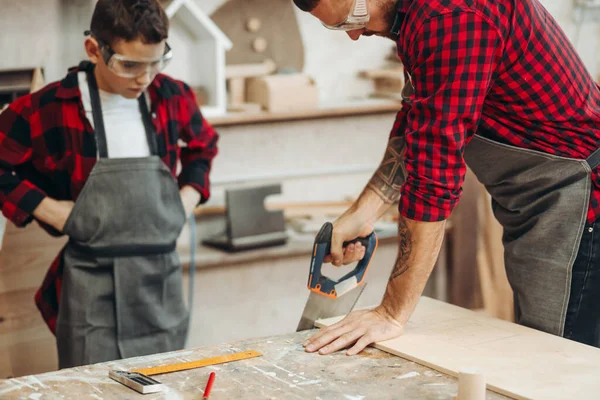 Papá y su hijo están trabajando en el taller. Están aserrando tablones de madera . — Foto de Stock