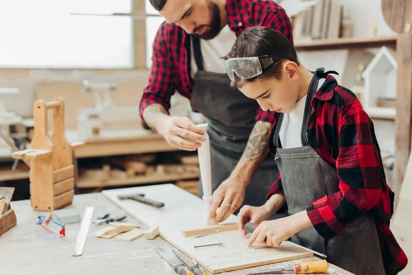 Malý chlapec pomáhá jeho tesař otec měření dřevěné prkno v dílně. — Stock fotografie