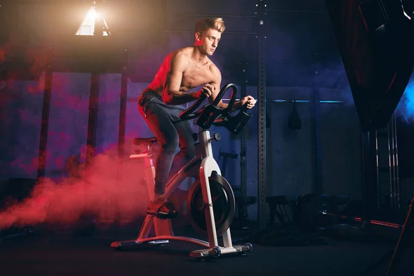 Desportista peito nu envolvido em um simulador de bicicleta no ginásio . — Fotografia de Stock