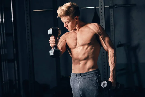 셔츠를 입지 않은 백인 남자가 어두운 체육관에서 덤벨 무게로 운동을 시작하다 — 스톡 사진