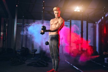 Kas gövdesi olan erkek antrenör, spor salonunda elinde dambıllar tutuyor.