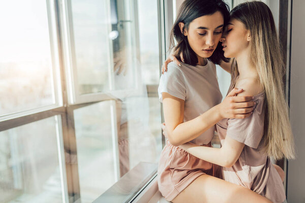 Two Lesbian women hugging together while standing near the window in bedroom.