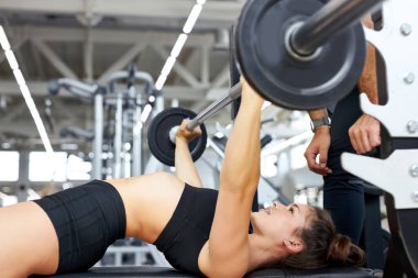 Genç bir kadın ve spor salonunda halter esneten kişisel antrenör.