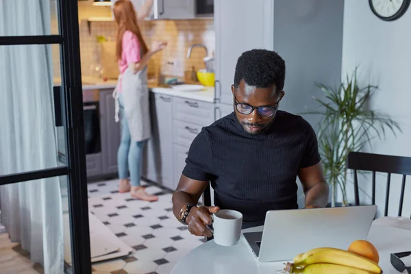 Confiado hombre africano trabajar en el ordenador portátil en casa, mientras que la esposa está ocupada en la cocina — Foto de Stock