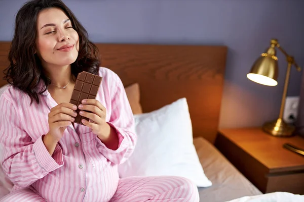 Smiling playful pregnant woman eating chocolate bar on bed — Stock Photo, Image