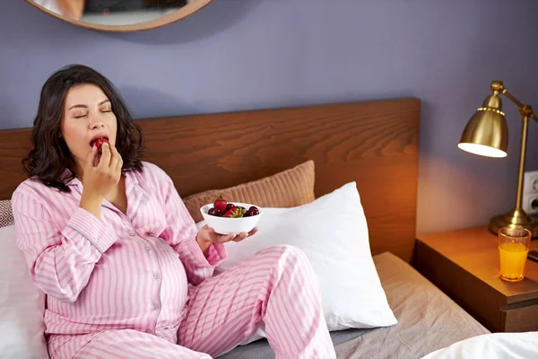 Pregnant woman eating strawberries sitting on bed at home — Stock Photo, Image